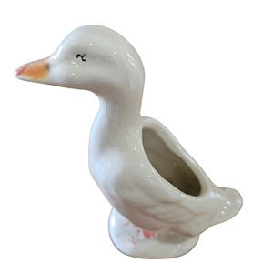 May include: A white ceramic duck-shaped planter with a glossy finish. The duck has a small orange beak, a painted black eye, and a hollow opening on its back for small items or plants. The base is rounded and features subtle orange accents.