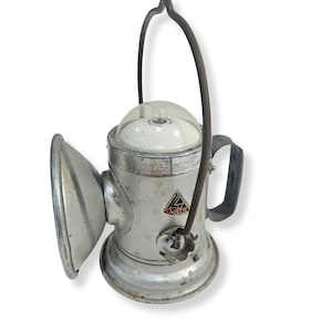 Peut inclure: Une lanterne en métal argenté vintage avec un réflecteur latéral, un dôme supérieur transparent et une poignée en fil métallique sombre. Le corps présente un logo triangulaire avec le texte Delta. La surface métallique montre de l'usure.