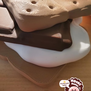 以下が含まれることがあります： スモアに似た、3Dプリントされた多層オブジェクト。上層と下層は、テクスチャ加工された表面と穴のあるタン色です。中間層は濃い茶色で、ベースは白です。木製の表面に置かれています。