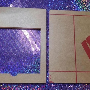 May include: Two pieces of light brown cardboard. One piece is a frame with a rectangular cutout. The other piece has red lines and the word MARGA in red. The background is a sparkly, iridescent purple.