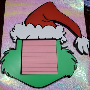 Op de afbeelding: Een grillige notitieblokhouder met kerstthema. Het ontwerp heeft een rode kerstmuts met witte rand, een groen gezicht en een roze notitieblokgedeelte met horizontale lijnen. De notitieblokhouder is gemaakt van een 3D-geprint materiaal.