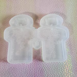 Puede incluir: Dos moldes de silicona transparentes y flexibles con forma de persona. Los moldes están conectados y tienen un ligero brillo. El fondo es una superficie texturizada e iridiscente con tonos rosas, verdes y amarillos.