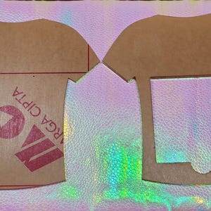 May include: Two brown acrylic t-shirt cutouts. One is a solid shape with red text and a logo that reads "ARGA CIPTA". The other is a t-shirt frame with a square and a rounded bottom cutout. The background is iridescent.