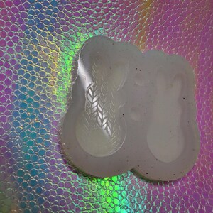 May include: A clear, flexible silicone mold with a textured surface. The mold features two spoon-shaped cavities, one with a leaf pattern. The background is a shimmering, iridescent surface.