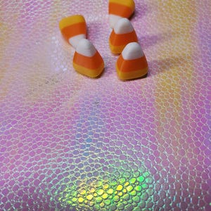 Könnte beinhalten: Mehrere Candy Corn-Bonbons liegen auf einer strukturierten, schillernden Oberfläche. Die Candy Corns haben das klassische dreifarbige Design aus Gelb, Orange und Weiß. Der Hintergrund hat ein rosa-violettes Farbschema.