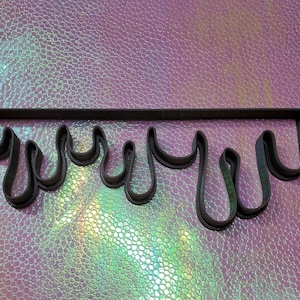 Puede incluir: Un cortador de galletas de metal gris oscuro con forma de líquido goteando. El cortador tiene una parte superior rectangular con varios recortes curvos en forma de lágrima colgando debajo. Sobre un fondo iridiscente y brillante.