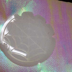 May include: A translucent, round coaster with a spiderweb design. The coaster has a scalloped edge and a small logo. The background is a shimmering, iridescent material with a textured pattern.