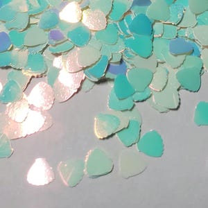 May include: A scattering of iridescent teal and white heart-shaped glitter. The glitter is scattered on a white surface.