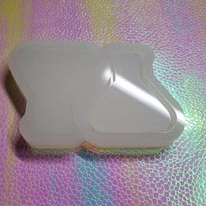 May include: A clear, two-part silicone mould for crafting, featuring shapes resembling laboratory beakers and flasks. The mould is translucent with small, scattered glitter particles. The background is a textured, iridescent surface with a rainbow sheen.