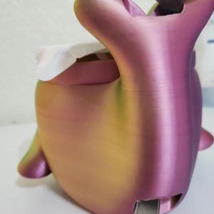 Może przedstawiać: 3D-drukowany pojemnik w kształcie wieloryba o gradientowym kolorze, przechodzącym od r&oacute;żowego do ż&oacute;łtego i zielonego. Posiada szczelinę na metalowy element i biały przedmiot w środku. Projekt obejmuje płetwy i ogon.