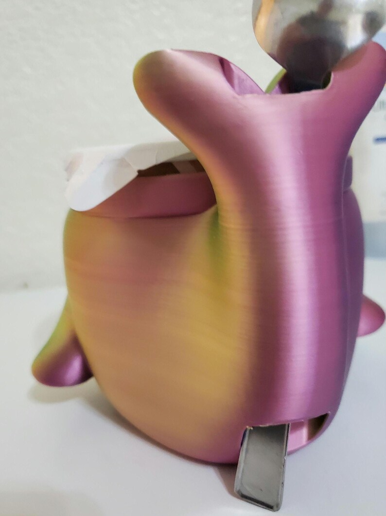 Może przedstawiać: 3D-drukowany pojemnik w kształcie wieloryba o gradientowym kolorze, przechodzącym od r&oacute;żowego do ż&oacute;łtego i zielonego. Posiada szczelinę na metalowy element i biały przedmiot w środku. Projekt obejmuje płetwy i ogon.