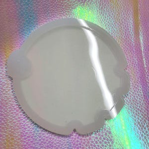 May include: A white silicone mold with a scalloped edge. The mold is designed to create a round, flat object, such as a coaster or a tray.