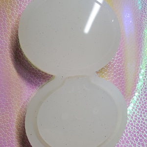 May include: A clear, open clam-shaped mould with a glittery, speckled appearance. The mould is made of a translucent material and is designed for crafting or casting. The two halves are connected at the base.