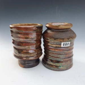 May include: Two handmade ceramic vessels with a textured, ridged design. The pottery features a blend of brown, copper, and iridescent hues. One vessel has a wider base, while the other is more cylindrical. The number "880" is on a white sticker.