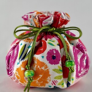 May include: A white drawstring bag with a floral print in shades of pink, orange, and green. The bag is tied with a green ribbon and has green beads on the ends.