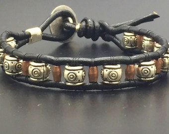 May include: A black leather bracelet with silver and copper beads. The bracelet has a simple knot closure.