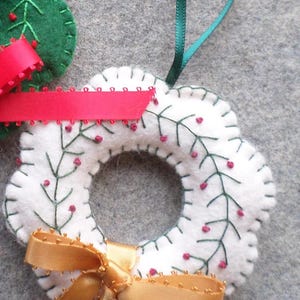 May include: A white felt wreath ornament with green embroidered branches and red berry accents. A gold ribbon bow and green ribbon loop for hanging are attached. A green felt tree ornament with a red ribbon is also visible.