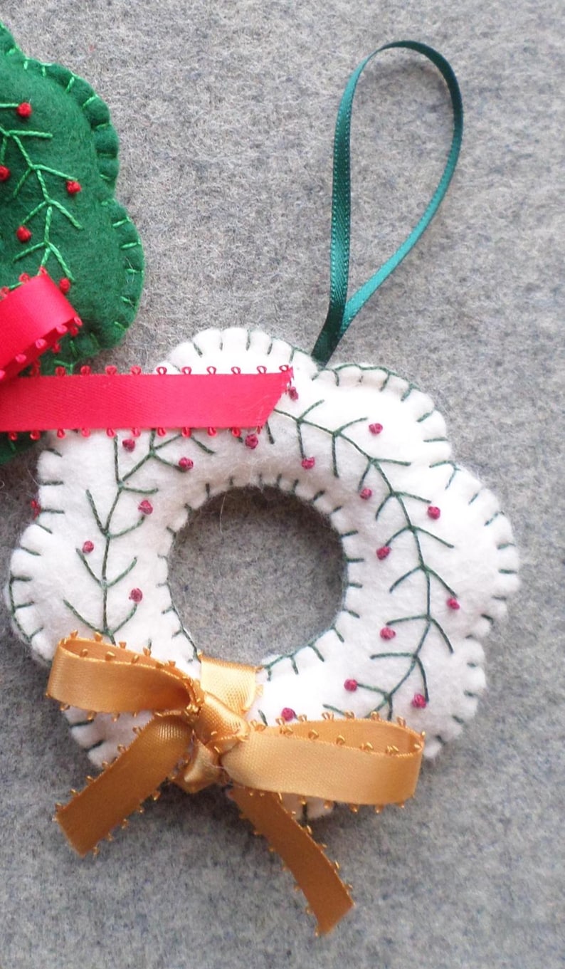 May include: A white felt wreath ornament with green embroidered branches and red berry accents. A gold ribbon bow and green ribbon loop for hanging are attached. A green felt tree ornament with a red ribbon is also visible.