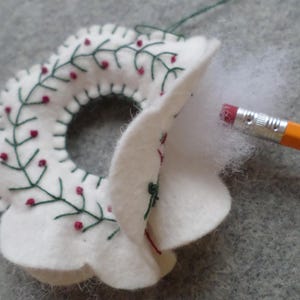 May include: A white felt Christmas wreath with green embroidered leaves and red berry accents. The wreath is being stuffed with white fiber fill using a yellow pencil. The wreath is on a gray surface.