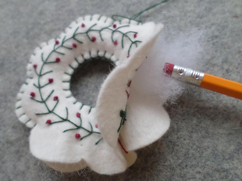 May include: A white felt Christmas wreath with green embroidered leaves and red berry accents. The wreath is being stuffed with white fiber fill using a yellow pencil. The wreath is on a gray surface.
