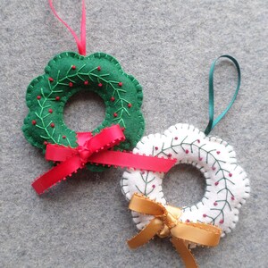 May include: Two felt Christmas wreath ornaments. One is green with red berries and a red ribbon. The other is white with red berries and a gold ribbon. Both have a loop for hanging.