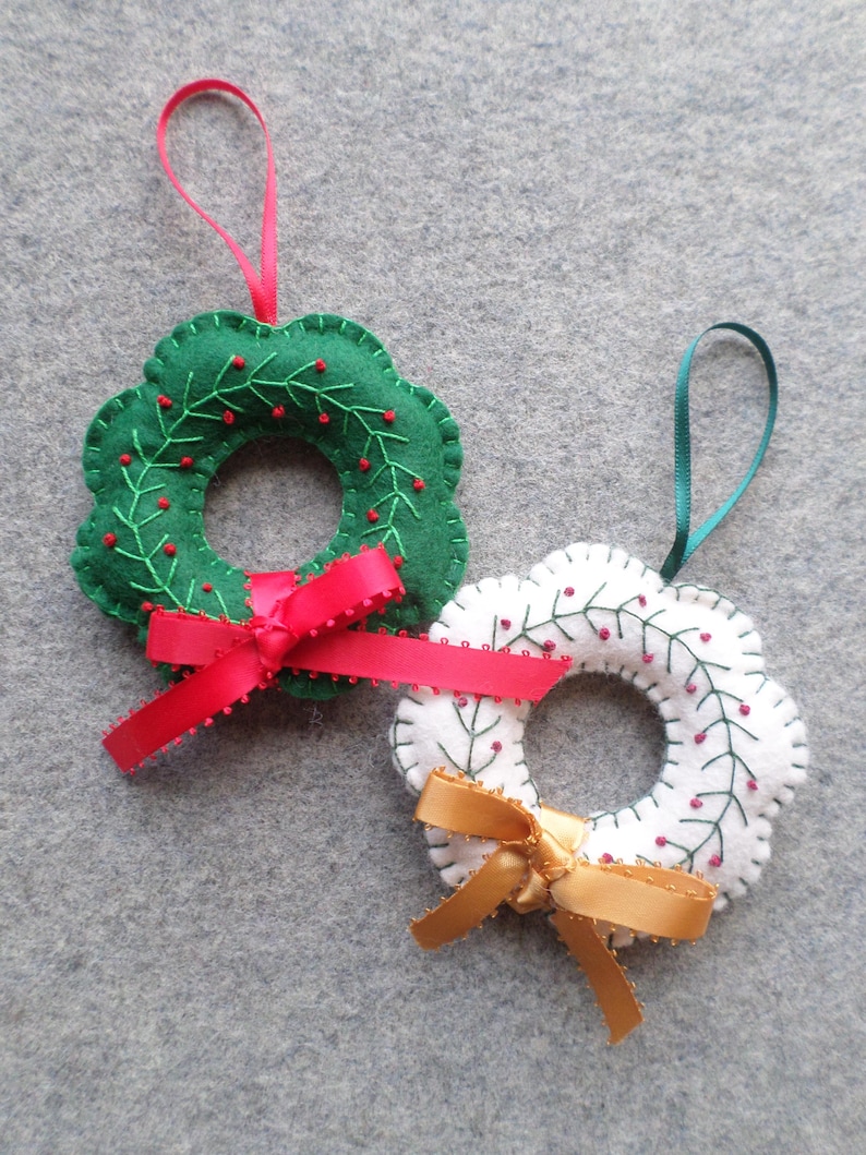 May include: Two felt Christmas wreath ornaments. One is green with red berries and a red ribbon. The other is white with red berries and a gold ribbon. Both have a loop for hanging.