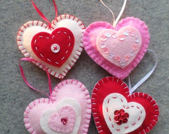 May include: Four handmade felt heart ornaments with hanging loops. One is white with a red heart and button, one is pink with a sequined heart, one is light pink with a white heart and beads, and one is red with a white heart and red sequins.