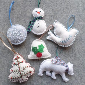 May include: A collection of handmade felt Christmas ornaments in various shapes, including a snowman, a dove, a bell, a tree, a polar bear, and a snowflake. Each ornament is white with colorful embroidered details and hanging ribbons. The text "Many More Designs Available" is at the top.