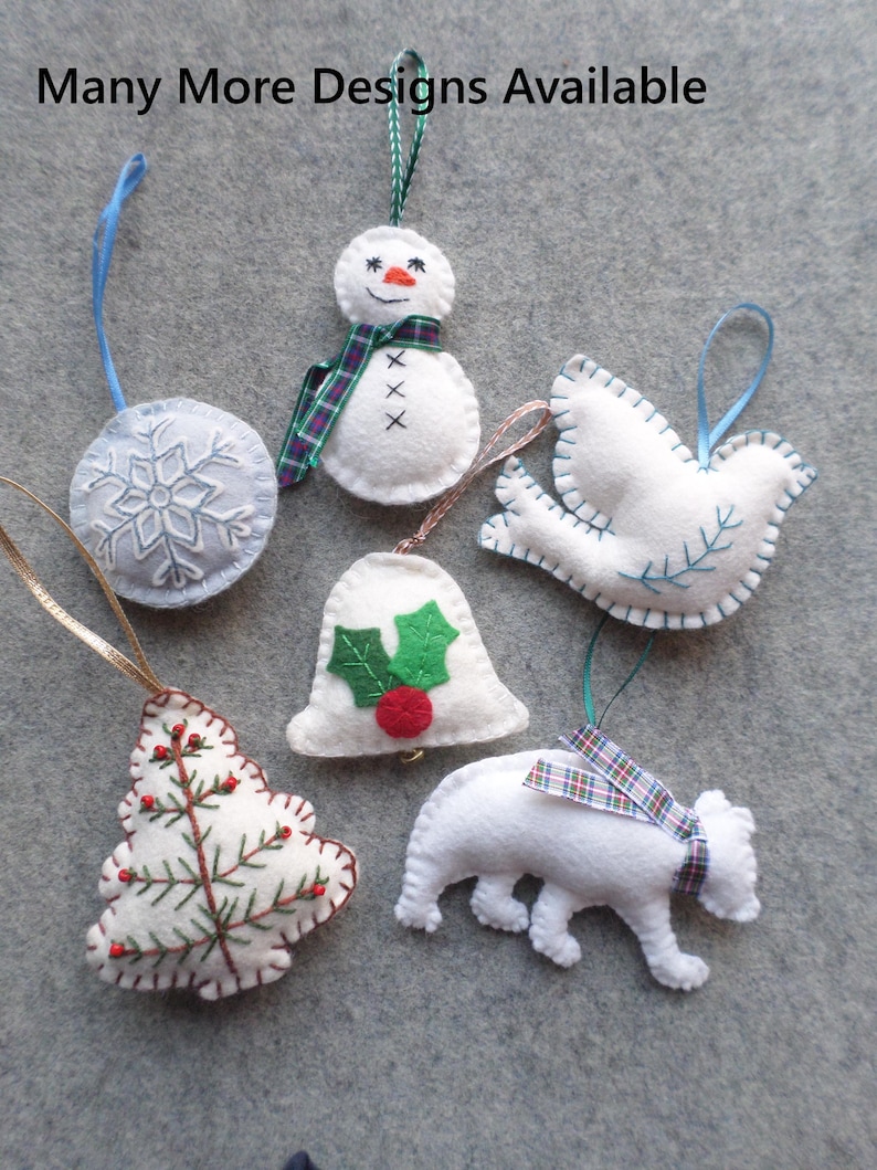 May include: A collection of handmade felt Christmas ornaments in various shapes, including a snowman, a dove, a bell, a tree, a polar bear, and a snowflake. Each ornament is white with colorful embroidered details and hanging ribbons. The text "Many More Designs Available" is at the top.