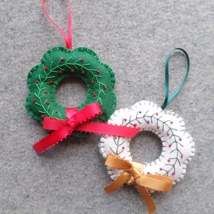 May include: Two felt Christmas wreath ornaments. One is green with red berries and a red ribbon, the other is white with red berries and a gold ribbon. Both have a loop for hanging.