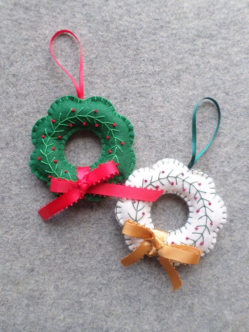 May include: Two felt Christmas wreath ornaments. One is green with red berries and a red ribbon, the other is white with red berries and a gold ribbon. Both have a loop for hanging.