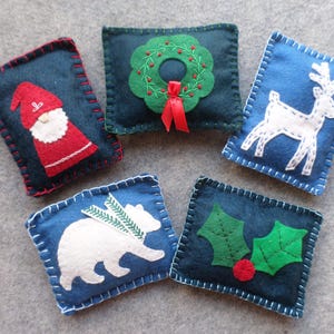 May include: Five handmade Christmas ornaments. Each ornament is a dark blue rectangle with a white stitched border. Designs include a Santa Claus, a wreath, a reindeer, a polar bear, and holly leaves.