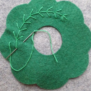May include: A green felt craft project with a scalloped edge and a central hole. Green embroidery thread is used to create a leaf-like design. A needle is threaded with green thread, showing the beginning of the embroidery process.