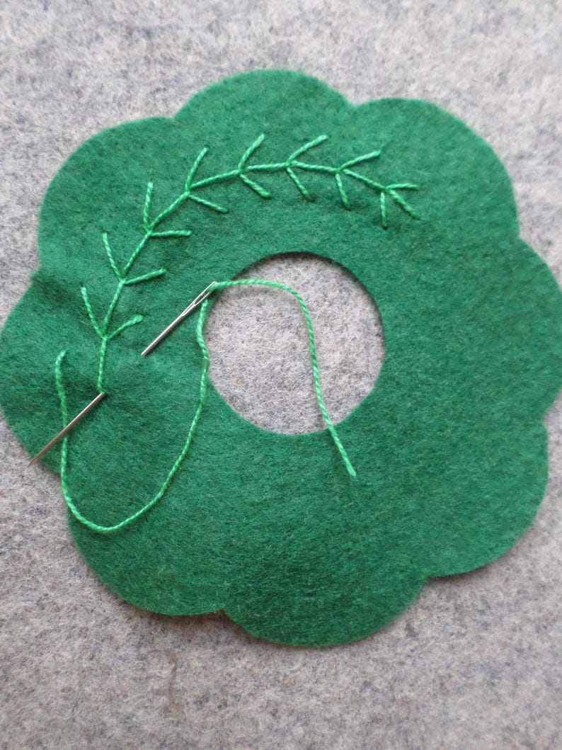 May include: A green felt craft project with a scalloped edge and a central hole. Green embroidery thread is used to create a leaf-like design. A needle is threaded with green thread, showing the beginning of the embroidery process.