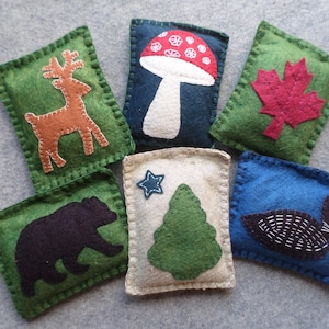 May include: Set of nine felt ornaments featuring various woodland creatures and nature scenes. The ornaments are green, blue, and beige with embroidered details. The ornaments include a deer, a mushroom, a maple leaf, a pine tree, and a loon.
