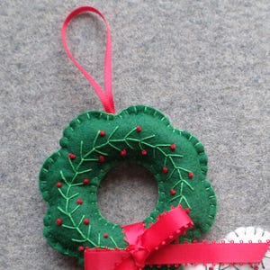 May include: A handmade green felt Christmas wreath ornament with red berry accents and a red satin ribbon bow. The wreath is embroidered with green vines and red berries. A pink ribbon loop is attached for hanging.