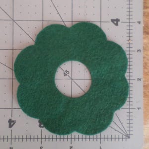 May include: A dark green felt flower shape with a circular hole in the center. The felt is on a gray cutting mat with inch measurements. The flower shape has a scalloped edge.