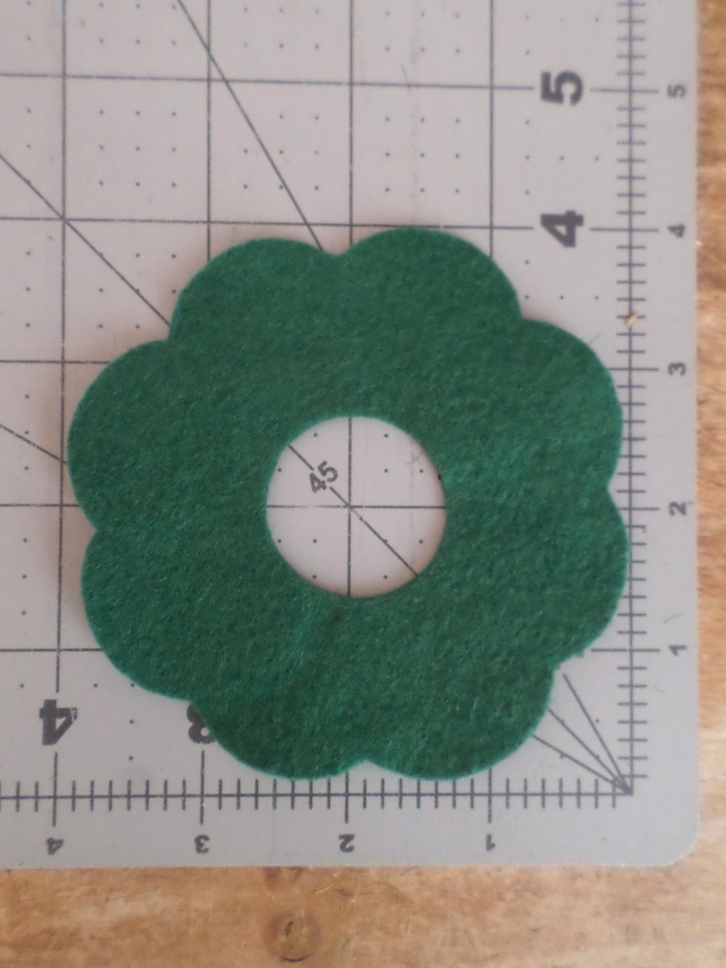 May include: A dark green felt flower shape with a circular hole in the center. The felt is on a gray cutting mat with inch measurements. The flower shape has a scalloped edge.