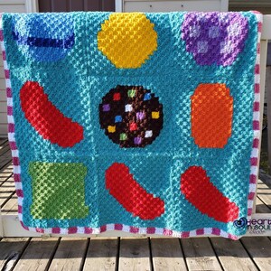 May include: A crocheted baby blanket with a colorful candy theme. The blanket features a variety of shapes, including circles, squares, and rectangles, in shades of red, orange, yellow, green, blue, and purple. The blanket is bordered with a pink and white striped trim.