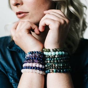 May include: A woman's wrists are adorned with a stack of colorful beaded bracelets. The bracelets are made of various gemstones, including amethyst, lapis lazuli, rose quartz, and jade. The bracelets are stacked on both wrists, creating a vibrant and eye-catching display.