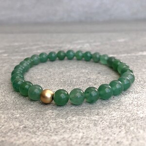 May include: A stretch bracelet composed of smooth, round green aventurine beads. The circular arrangement features a single, polished gold-tone metal bead positioned as a focal point among the uniform green gemstone beads.