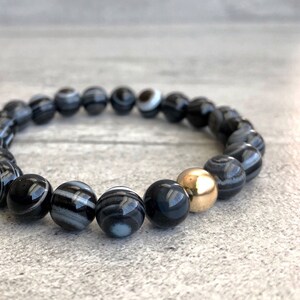 May include: Black and white agate gemstone bracelet with a gold bead accent. The bracelet is made of round beads and is approximately 7 inches in length.