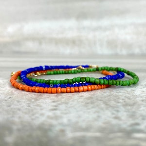 May include: A layered bracelet made of small, colorful beads. The bracelet is made of three strands of beads: orange, green, and blue.