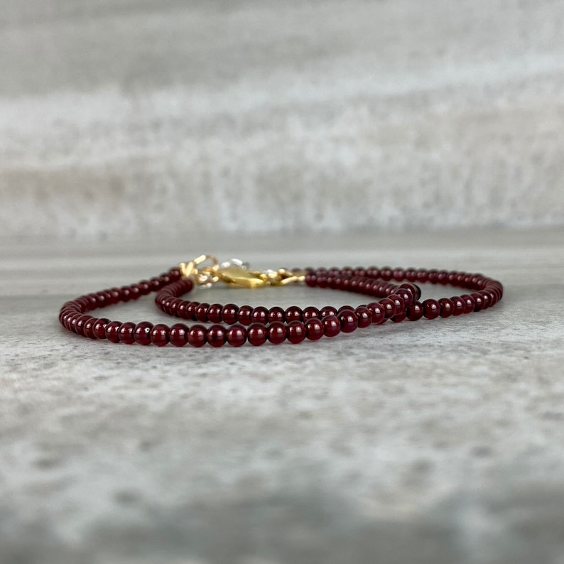 May include: A delicate necklace made of small, dark red garnet beads. The necklace is strung on a gold chain and has a gold clasp.