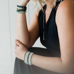 May include: A stack of four beaded bracelets in shades of blue and green worn on the upper arm, and a stack of four light green and white beaded bracelets worn on the lower arm, paired with a silver chain necklace featuring a small pendant.
