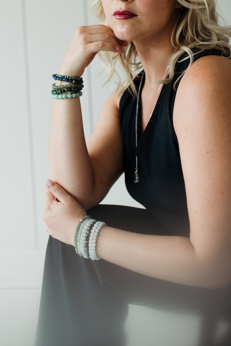 May include: A stack of four beaded bracelets in shades of blue and green worn on the upper arm, and a stack of four light green and white beaded bracelets worn on the lower arm, paired with a silver chain necklace featuring a small pendant.