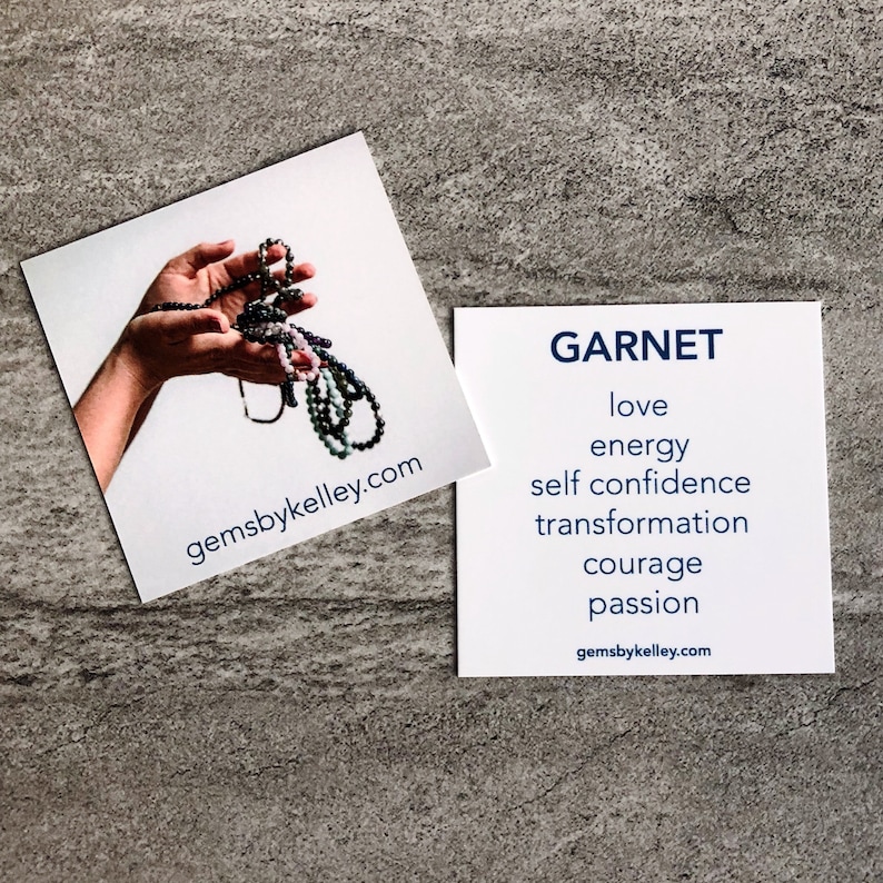 May include: A white card with the word "GARNET" in blue text. Below the word "GARNET" are the words "love", "energy", "self confidence", "transformation", "courage", and "passion" in blue text. The words "gemsbykelley.com" are in blue text at the bottom of the card.