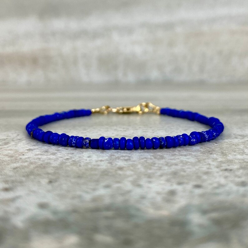 May include: A delicate blue beaded bracelet with a gold clasp. The bracelet is made of small, round beads that are strung together on a thin cord.