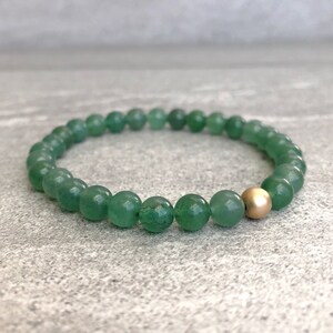 May include: A beaded bracelet featuring numerous small, round, translucent green stone beads arranged in a circular pattern, accented by a single, smooth, matte-finish gold-colored metal bead positioned prominently along the strand.