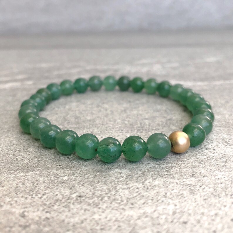 May include: A beaded bracelet featuring numerous small, round, translucent green stone beads arranged in a circular pattern, accented by a single, smooth, matte-finish gold-colored metal bead positioned prominently along the strand.